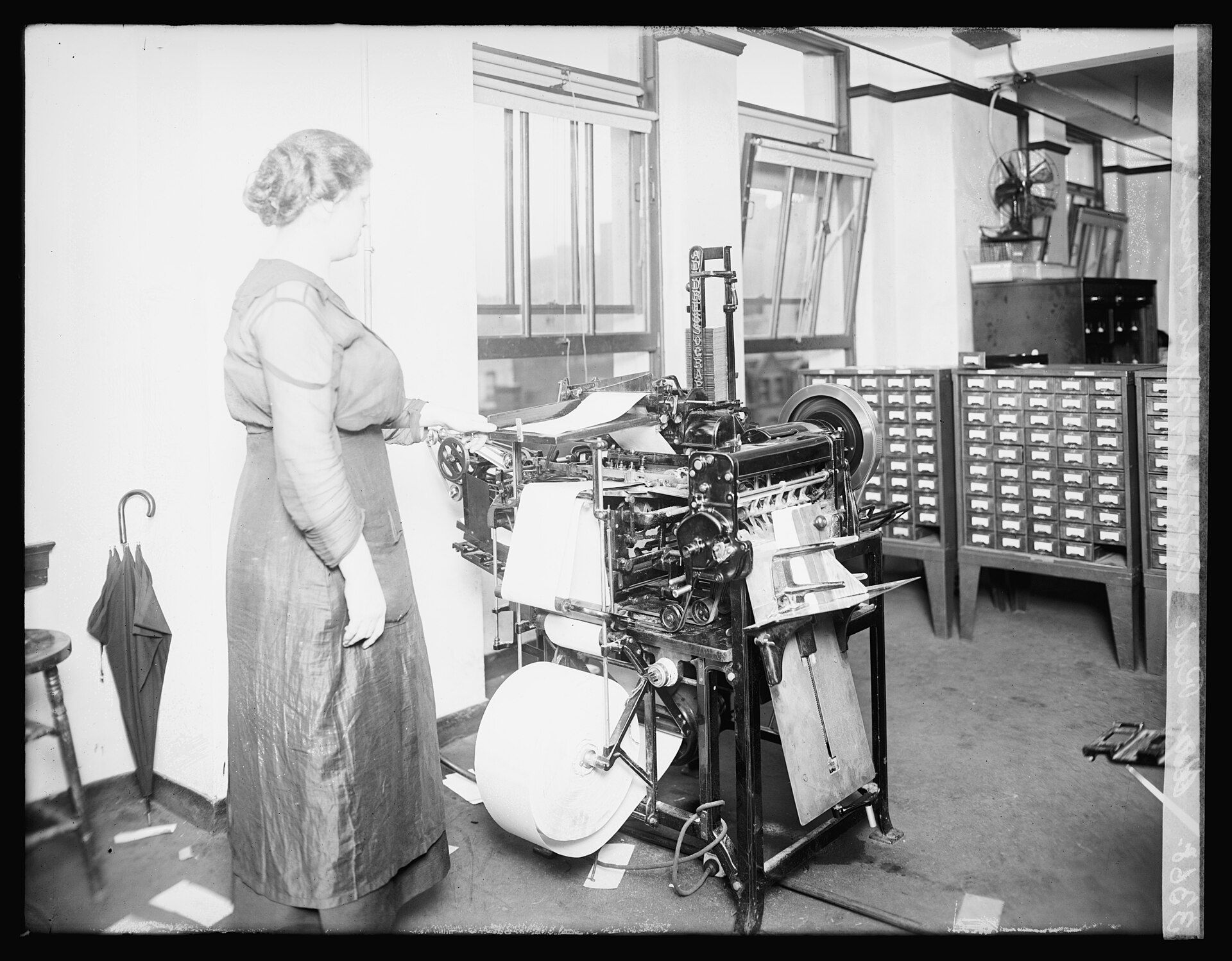 A woman operating an Addressograph mailing-list printer beside rows of card-file cabinets in an office
