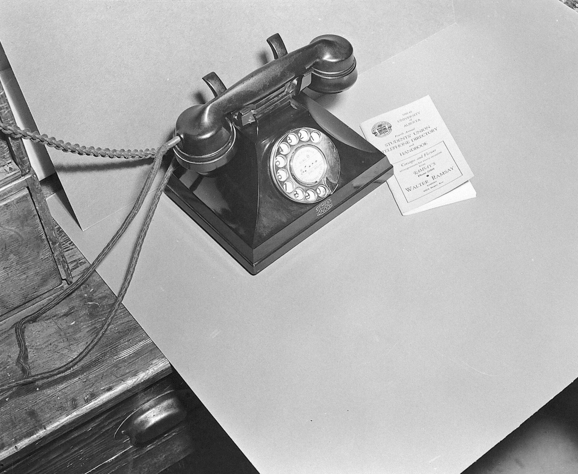 A black rotary desk telephone beside a small 1943 student telephone directory
